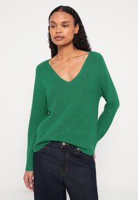 Žalias raštuotas mezginys su V formos iškirpte, ilgomis rankovėmis ir mažo logotipo detale. Dėvimas su tamsiai mėlynomis džinsomis. Minkšta tekstūra.