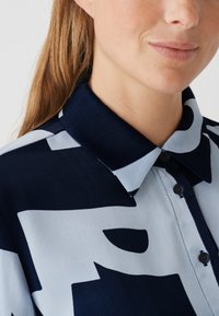 Hemd mit Knöpfen in Marineblau und Hellblau, mit geometrischem Muster. Weicher Stoff, Spitzenkragen und kleine Knöpfe als Detail.