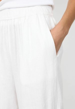 Desna ruka smještena u bočni džep širokih bijelih hlača s elastičnim pojasom, nošene s bijelom majicom.