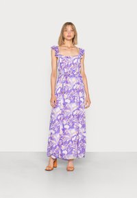 Vestido maxi em padrão floral roxo com alças com babados e decote quadrado. Apresenta uma cintura ajustada e saia fluida. Sapatos de salto alto laranja.