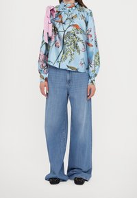 Donna che indossa una blusa floreale blu con un fiocco rosa sulla spalla, jeans larghi azzurri chiari e scarpe nere con borchie.