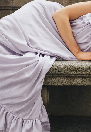 Osoba v svetlofialových šatách ležiaca na boku na kamennej lavičke s jednou rukou opretou o povrch.