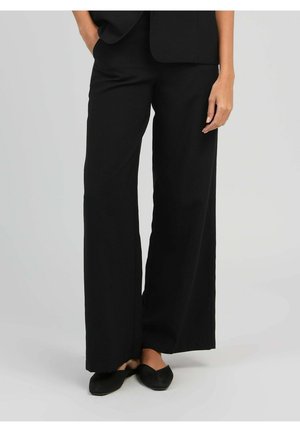 Femme portant un pantalon noir à jambes larges et des chaussures plates noires, debout avec une main dans la poche et l'autre détendue le long du corps.