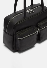 Sac en cuir noir avec deux poches zippées externes, doubles poignées supérieures et une texture lisse. Fermetures éclair en métal et détail de logo embossé visibles.