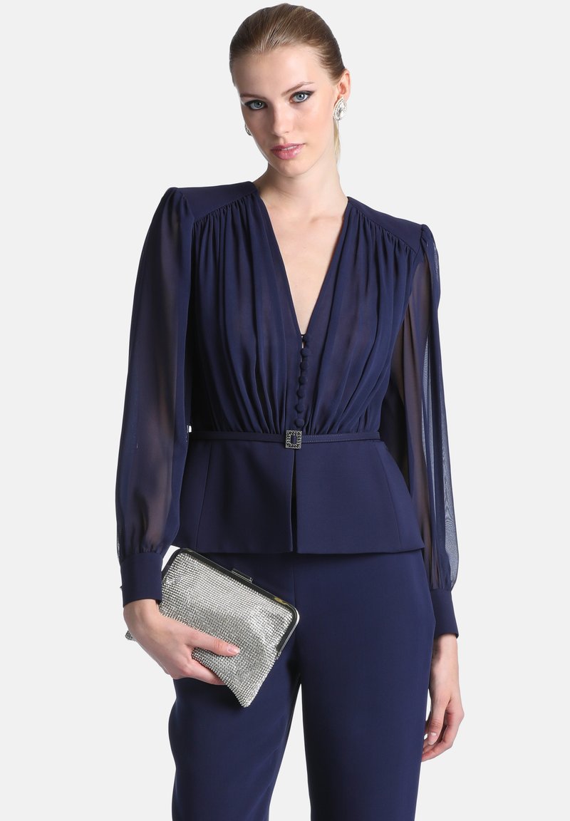 Jeune femme aux cheveux clairs portant une blouse bleu marine à manches longues et un pantalon, tenant une pochette argentée texturée.