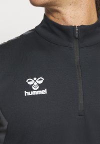 Top sportivo nero con zip, realizzato in un tessuto testurizzato, logo bianco e accenti grigi sulle spalle. Collo liscio e design aderente.