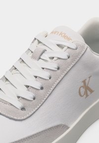 Baskets Calvin Klein blanches et beige montrant les lacets, textures en daim et tissu, avec le logo "cK" sur le côté et le nom de la marque sur la languette.