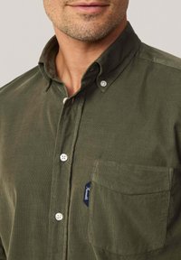 Camisa de pana de color verde oliva con cuello abotonado, que presenta botones blancos y un bolsillo en el pecho con una etiqueta azul. Se aprecia un detalle de tela texturizada.