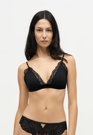 Femme aux longs cheveux foncés, portant un soutien-gorge en dentelle noire et une culotte assortie, debout devant un fond clair uni.