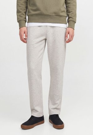 Homme portant un pantalon de jogging gris clair, un sweat-shirt vert olive, un sous-vêtement blanc et des baskets noires avec des semelles marron, debout sur un fond blanc.