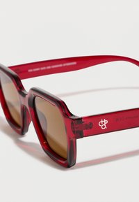 Κόκκινα διαφανή γυαλιά ηλίου με τετράγωνο σκελετό και καφετί φακούς που ακουμπούν σε λευκή επιφάνεια.