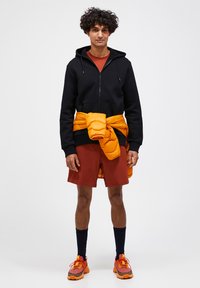 Sort hættejakke med lynlås, rustfarvet t-shirt, orange puffjakke bundet om livet, maroon shorts, navy strømper og orange og grå sneakers.