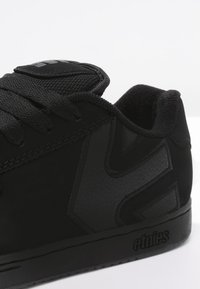 Schwarzer Sportschuh aus Wildleder und synthetischem Material. Verfügt über schwarze Schnürsenkel, einen gepolsterten Schaft und ein geprägtes Logo an der Seite. Glatte Textur.
