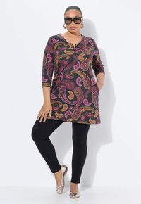 Schwarze Tunika mit lebhaftem Paisleymuster in Pink und Gold, kombiniert mit schwarzen Leggings und metallischen Ballerinas. Modell trägt große Sonnenbrille.