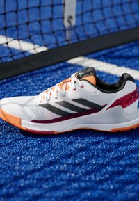 Witte sportieve schoen met zwarte strepen, oranje accenten en een gestructureerde zool. Heeft een bovenwerk van mesh en een gevoerde kraag voor comfort.