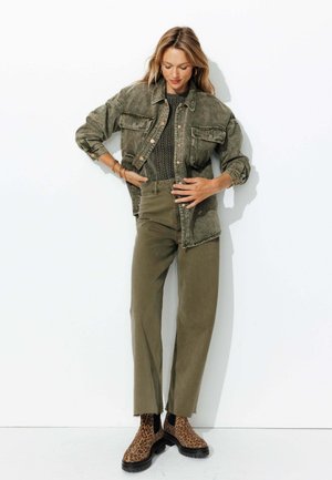 Femme portant une veste vert olive, un top en crochet, un pantalon assorti et des bottes imprimé léopard, se tenant devant un fond blanc.