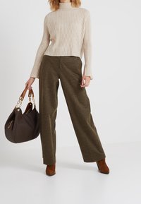 Suéter de punto beige con cuello alto, combinado con pantalones anchos en verde olivo. Botas de tacón marrones y bolso oscuro con correa de cadena.