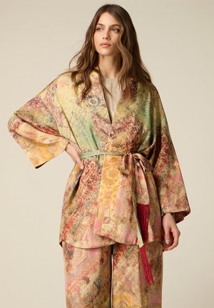 Donna che indossa una giacca stile kimono larga e fantasia e pantaloni a gamba larga abbinati con cintura decorativa e nappina, in piedi su sfondo semplice.