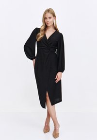 Schwarzes Wickelkleid mit langen Puffärmeln, einem gedrehten Vorderdesign und einem hohen Schlitz. Der Stoff hat einen dezenten Schimmer und eine glatte Textur.