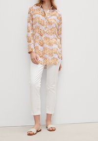 Camicia a maniche lunghe bianca con un motivo a onde arancioni, chiusura con bottoni e una vestibilità rilassata, abbinata a pantaloni bianchi e sandali leggeri.