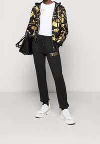 Haut à capuche noir avec imprimé chaîne dorée, t-shirt blanc avec logo, pantalon de survêtement noir avec logo doré, et baskets blanches épaisses. Sac à main noir.