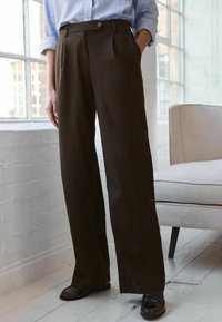 Bruine, wijdvallende broek gemaakt van een zachte stof, met voorplooien en een knoopsluiting. Gecombineerd met zwarte schoenen, tegen een lichte achtergrond.
