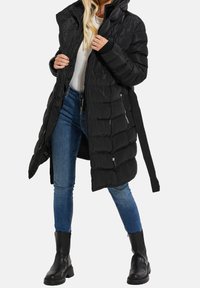 Mujer con un abrigo acolchado negro con capucha, jeans ajustados azules, botas negras y una blusa blanca, ajustando la parte delantera del abrigo.