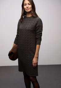 Sweterkowa sukienka do kolan w ciemnobrązowym kolorze z teksturowanym wzorem, długimi rękawami i okrągłym dekoltem. Modelka trzyma brązową kopertówkę.