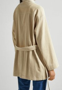 Giacca di velluto a coste beige con righe verticali, maniche lunghe e cintura annodata in vita. Presenta una texture morbida e una vestibilità rilassata.