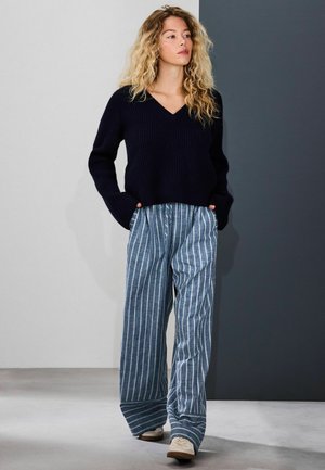 Marineblauer Rippstrickpullover mit V-Ausschnitt und langen Ärmeln, kombiniert mit blau-weiß gestreiften Hosen mit weitem Bein; legere Schuhe runden das Outfit ab.