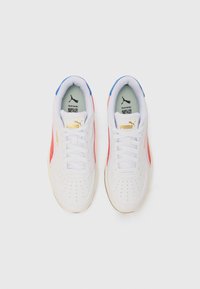 Scarpe da ginnastica bianche con punta perforata, caratterizzate da accenti rossi e blu, lacci bianchi e un logo dorato sul lato. Texture liscia.