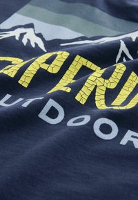 Marinblått tyg med ett texturerat gult "OUTDOORS" grafiskt tryck och bergdesign i nyanser av blått och grönt, med ett sprucket tryckseffekt.
