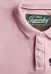 Hellrosa Polohemd mit zwei weißen Knöpfen, umgeklapptem Kragen und sichtbarem "Superdry" Markenlabel innen am Kragen.