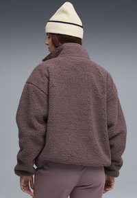 Maglione in pile marrone con collo alto e vestibilità rilassata. La texture è morbida e shaggy, abbinata a un berretto lavorato a maglia crema con una striscia scura.