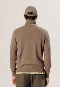 Bruine gebreide trui met een hoge kraag, aansluitende mouwen en een geribbelde zoom. Draagt een olijfgroene shirt en een grijze pet.