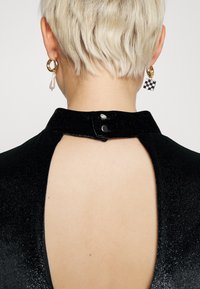 Robe en tissu noir scintillant avec un col montant, dotée d'une ouverture en forme de trou de serrure dans le dos et de boutons-pression au niveau du cou. Boucles d'oreilles anneaux dorés et perles.