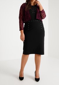 Černé šaty s přiléhavým střihem, zdobené dekorativními cvočky a hlubokým výstřihem, doplněné krátkým vínově červeným krajkovým sáčkem. Černé podpatky.