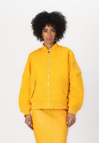 Giacca bomber gialla con polsini a costine, chiusura con zip e tasche laterali. Tessuto morbido, foderata imbottita, presenta una tasca con zip sulla manica.