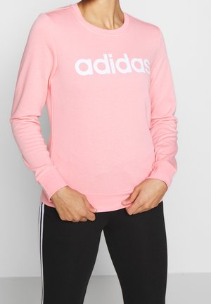 Osoba ubrana w różową bluzę Adidas z długimi rękawami i czarne spodnie z białymi paskami, ręce spoczywają blisko pasa.