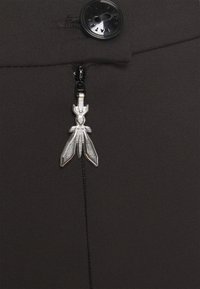 Dunkler Stoff mit einem Reißverschluss, der einen silbernen Ziehgriff in Form eines stilisierten Insekts aufweist, ergänzt durch einen runden schwarzen Knopf darüber.