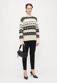 Knitpullover in Creme mit schwarzen geometrischen Mustern, kombiniert mit schmalen schwarzen Hosen und hohen schwarzen Schuhen, während sie eine kleine schwarze Tasche hält.