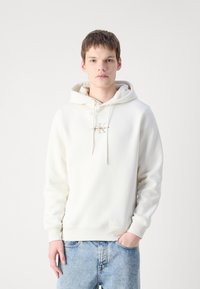 Sudadera blanca de algodón con un bolsillo frontal, que presenta un logo dorado de Calvin Klein. El diseño incluye una capucha con cordones y puños acanalados.