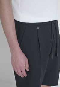 Pantaloni neri con pieghe e vita con coulisse, caratterizzati da un accento metallico. Abbinati a una maglietta bianca a maniche corte, visti di lato.