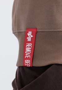 Sudadera marrón con puños acanalados y una etiqueta roja que dice "REMOVE BEF" en texto blanco, con una textura suave y un diseño estructurado.