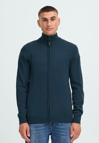 Ung mann står, iført en mørkeblå zip-up genser med ribbestrikkede mansjetter og krage, kombinert med blå jeans, mot en ensfarget bakgrunn.