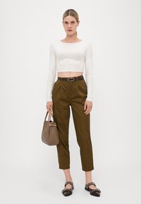 Bela dolgi rokavi crop top, olivni hlače s prilagojenim krojem, rjav pas in temni sandali z dekorativnimi elementi. Rjava torbica ob strani.