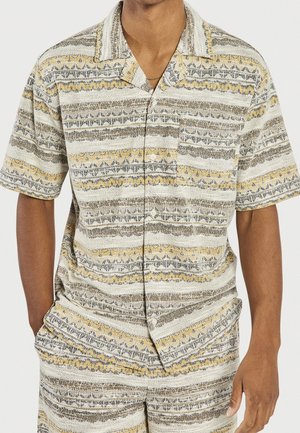 Mand iført en kortærmet skjorte med knapper og matchende shorts med horisontale striber i beige, gul og grå mønstre.