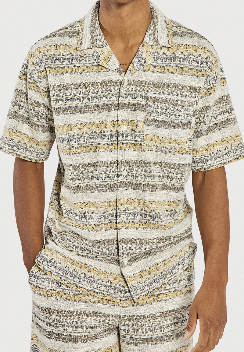 Homme portant une chemise à manches courtes boutonnée et un short assorti avec des rayures horizontales en beige, jaune et gris.