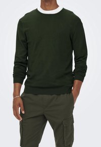 Pull vert foncé avec un col rond blanc, poignets et ourlet côtelés. Porté avec un pantalon cargo assorti, doté de poches latérales.