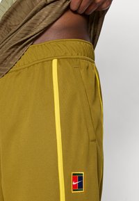 Ναϊκ ελαιόχρωμα σορτς με κίτρινες λωρίδες στο πλάι, ελαστική μέση και μια μικρή πολύχρωμη ετικέτα λογοτύπου στο κατώτερο μέρος.